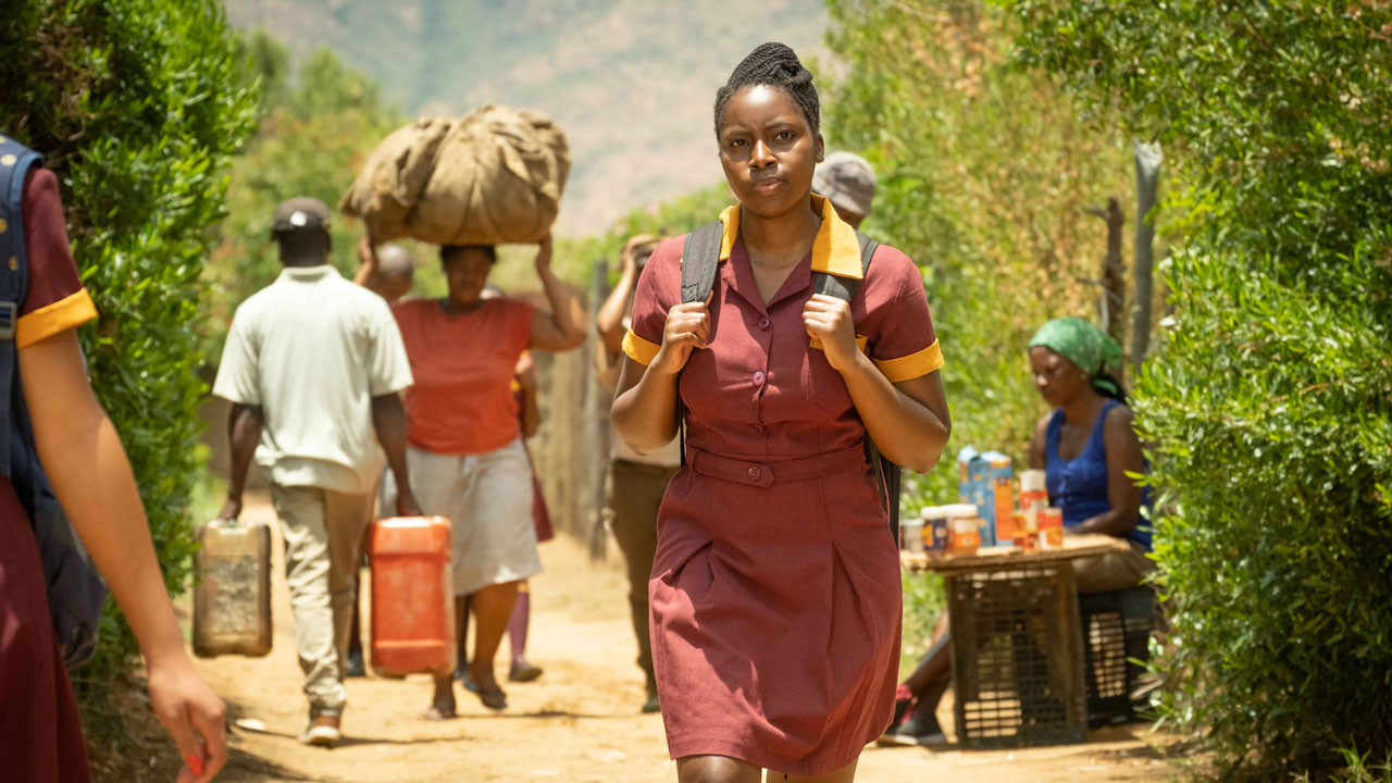 Girl in school uniform walking, with vendor and other people carry load in the background