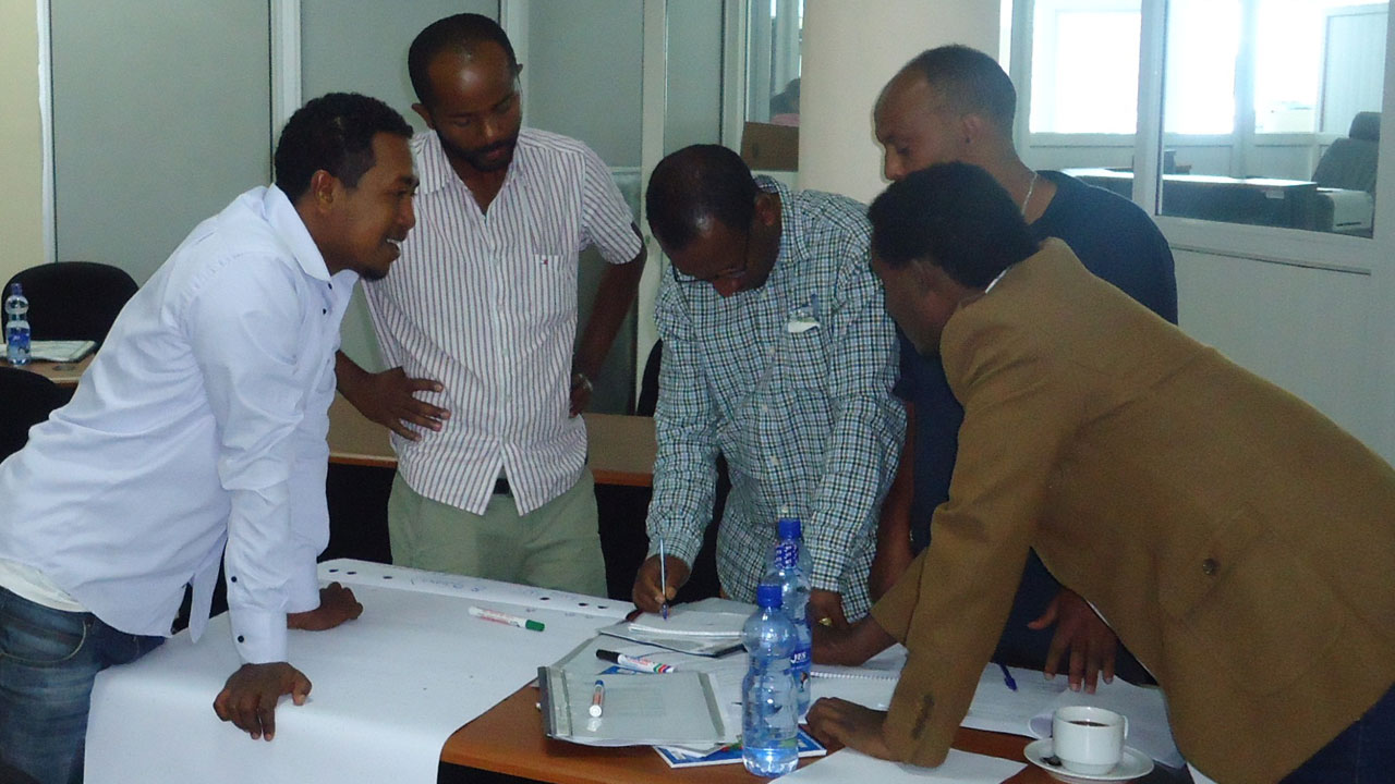 Five males who are in a workshop activity with one writing in a book while the others look on