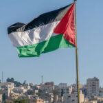 The large Palestinian flag is waiving above the city with tall buildings in the background