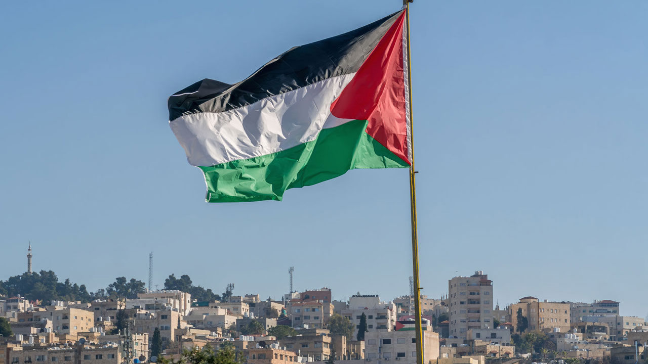 The large Palestinian flag is waiving above the city with tall buildings in the background