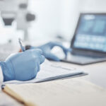 Gloved hands writing notes on a notebook with laptop findings and microscope in the background