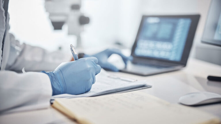 Gloved hands writing notes on a notebook with laptop findings and microscope in the background