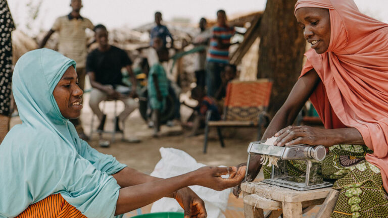 A woman hands another woman food as she puts it into a food processor machine