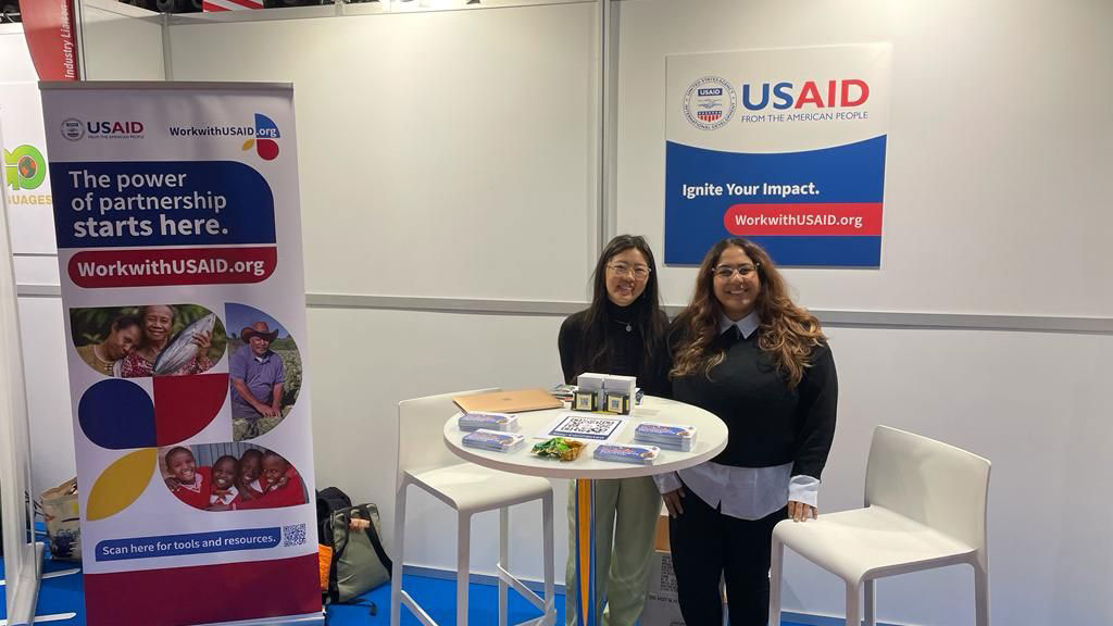 Two people standing around a table with USAID posters in their surroundings