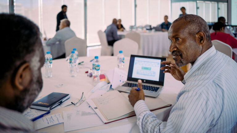 Two men at a table engaged in discussion, laptop in front of them